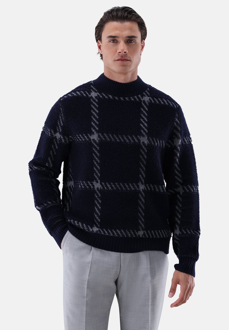 Marineblauer Pullover mit strukturiertem Strick, der ein graues Karomuster zeigt. Hoher Kragen und gerippte Bündchen, kombiniert mit hellgrauen Hosen.