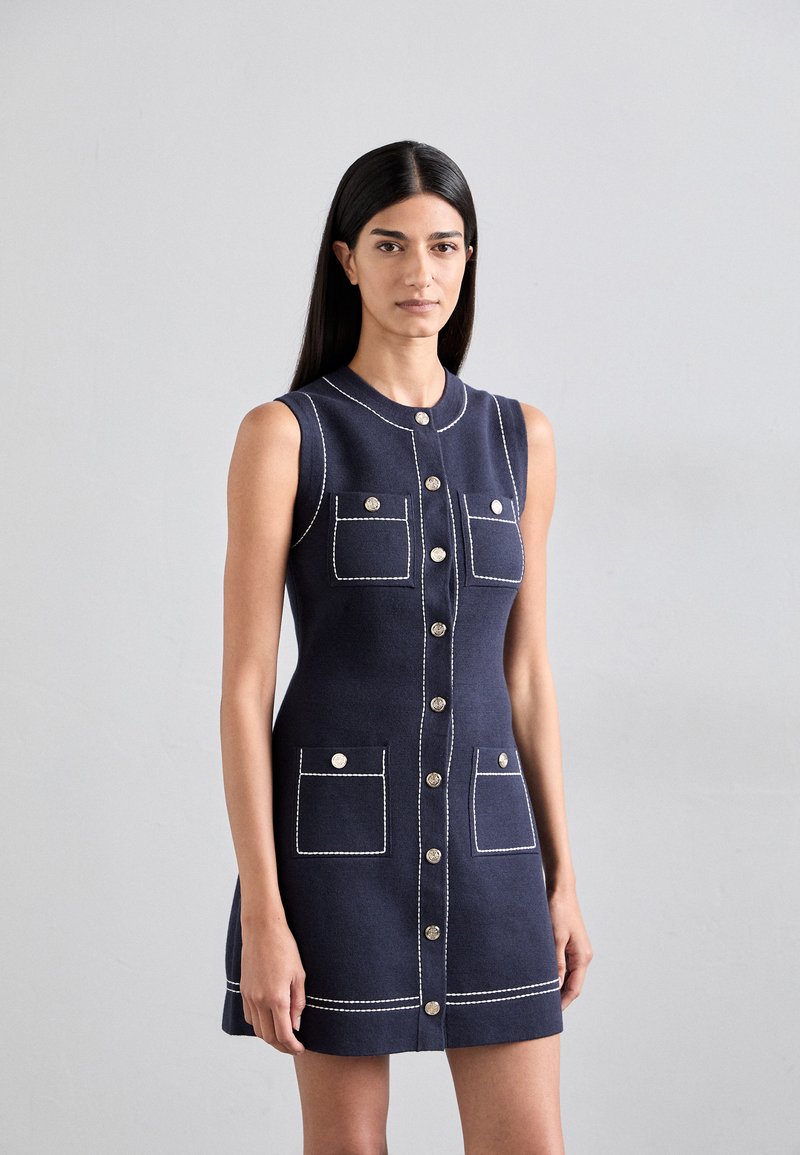 Marineblauwe mouwloze jurk met zilveren knopen, twee voorkant zakken, witte stikseldetails en een getailleerd silhouet. Zachte stoftextuur.