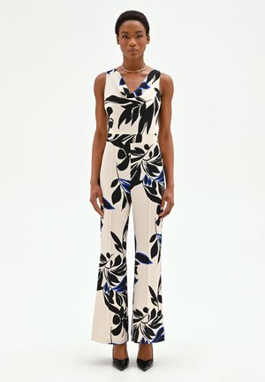 Donna che indossa una tuta senza maniche color crema con grandi motivi floreali neri e blu, abbinata a tacchi neri a punta, su sfondo bianco.