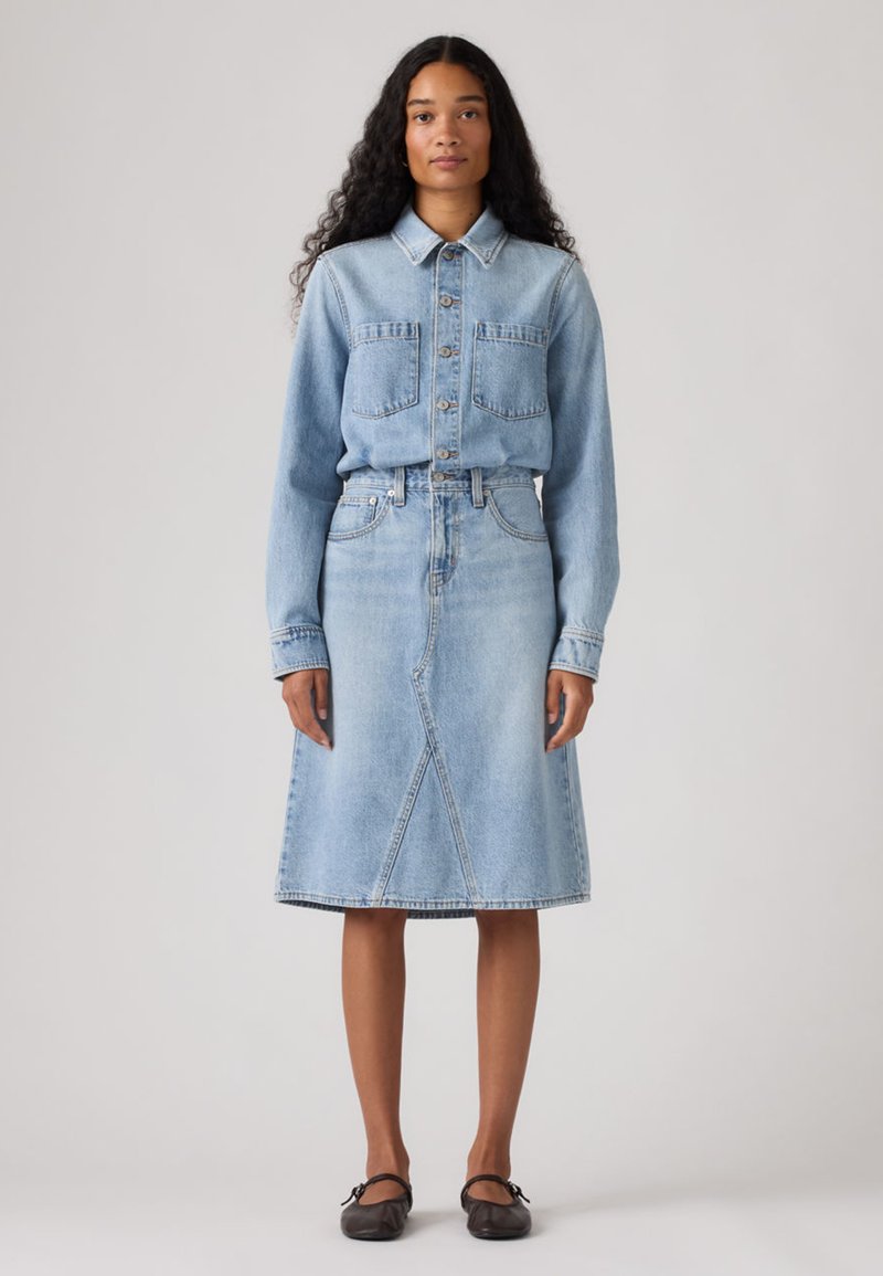 Ανοιχτό μπλε πουκάμισο τζιν με κουμπιά και δύο τσέπες στο στήθος, σε συνδυασμό με φούστα τζιν μήκους μέχρι το γόνατο, στο ίδιο χρώμα. Ομαλή υφή, κλασικός σχεδιασμός.