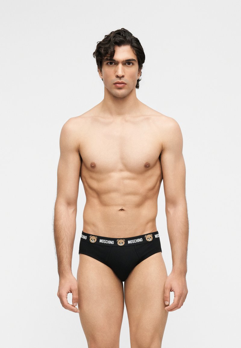 Άνδρας χωρίς πουκάμισο με κοντά κυματιστά μαλλιά που φοράει μαύρα εσώρουχα Moschino με λογότυπο αρκουδάκι στη μέση, στέκεται μπροστά σε απλό φόντο.