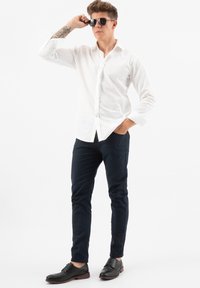 Camicia bianca con bottoni, pantaloni blu navy e scarpe nere. Modello che indossa occhiali da sole, posando con la mano sulla fronte. Tessuto liscio, vestibilità su misura.