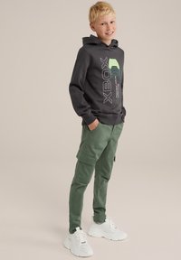 Grijze hoodie met groene en blauwe Xbox-afbeelding, gecombineerd met olijfgroene cargo-broek met zakken en witte chunky sneakers.