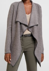 Šedý pletený cardigan s herringbone vzorem, širokým splývavým límcem, dlouhými rukávy a asymetrickým lemem. Nositý přes béžový top a zelené kalhoty.