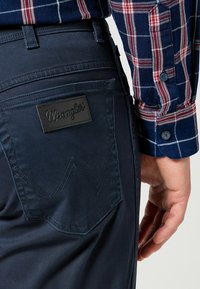 Pantalones Wrangler azul marino con una etiqueta de cuero y un bolsillo trasero en ángulo; llevados con una camisa de manga larga a cuadros en azul, rojo y blanco.