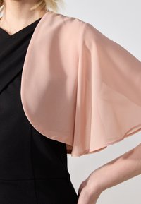 Zbliżenie na osobę ubraną w przezroczysty, zwiewny jasnoróżowy szal narzucony na dopasowany czarny strój, z ręką na biodrze na jednolitym tle.