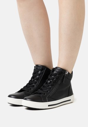 Sneakers nere in pelle alta con lacci e cerniera laterale indossate su gambe nude su sfondo bianco semplice.