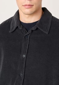 Muškarac obučen u tamnosivu kordiranu košulju na kopčanje preko crnog džempera okruglog vrata, prikazan vratni izrez i gornji dio prsa.