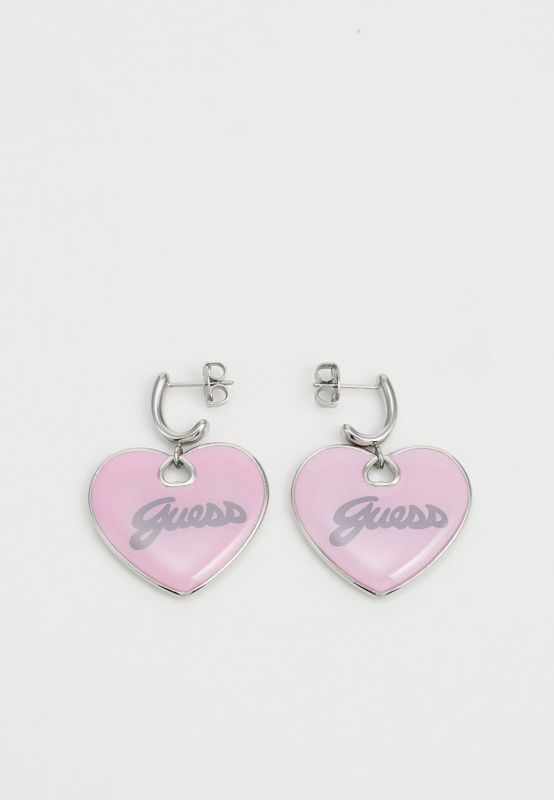 Orecchini a forma di cuore in smalto lucido rosa, con il logo "Guess" in grigio, dotati di hardware in metallo argentato e chiusura a gancio.