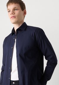 Cămașă albastră marin, cu guler clasic, dintr-un material neted, cu mâneci lungi și nasturi gri pe față.