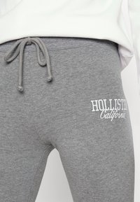 Grå träningsoverallsbyxor med elastisk midja och dragsko; har en vit broderad "Hollister California"-logotyp på vänster lår.