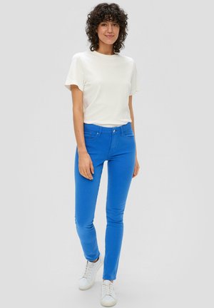 Jean slim fit bleu vif avec cinq poches, associé à une chemise blanche à manches courtes unie et des baskets blanches. Texture lisse, design décontracté.