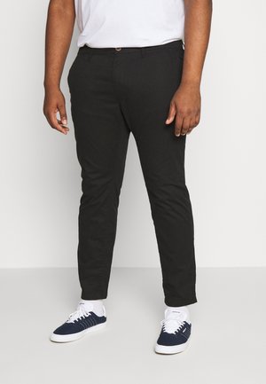 Mężczyzna ubrany w czarne spodnie slim-fit, białe skarpetki za kostkę, granatowo-białe buty sportowe Adidas oraz białą koszulkę, stojący na szarym tle.