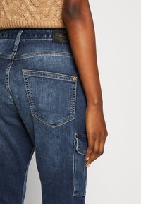 Mörk denim cargo-byxor har en texturerad finish, flera fickor och en åtsittande design. Tyget är robust med synliga sömmar som accent.