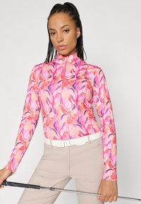 Skaidra rozā garās piedurknes golfa krekls ar ziedu rakstu rozā un purpursarkanā toņos. Tam ir augsts apkakle un pus-izsist aizdare.