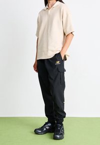 Person standing with hands in pockets wearing beige polo, black New Balance cargo pants, and black athletic shoes on green floor.