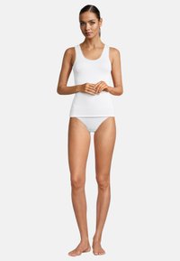 Top bianco senza maniche con bordo in pizzo e bikini bianco abbinato. Design semplice, forma aderente e texture liscia. Modella in piedi a piedi nudi con le mani incrociate.