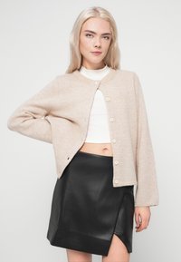 Svetlo bež pleteni kardigan s okruglim izrezom, pet dugmadi i dugim rukavima, stiliziran preko bele majice i crne kožne suknje.