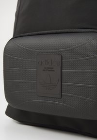 Fekete hátizsák, texturált szövetből készült, középen kiemelkedő fekete gumiból készült Adidas logóval, amely háromcsíkkal és sima részletekkel van ellátva.