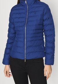 Sininen puhaltava takki, jossa on korkea kaulus, tikattu muotoilu, etuvetoketju ja logon korostus. Sileä, kevyt kangas ja hienostunut istuvuus.