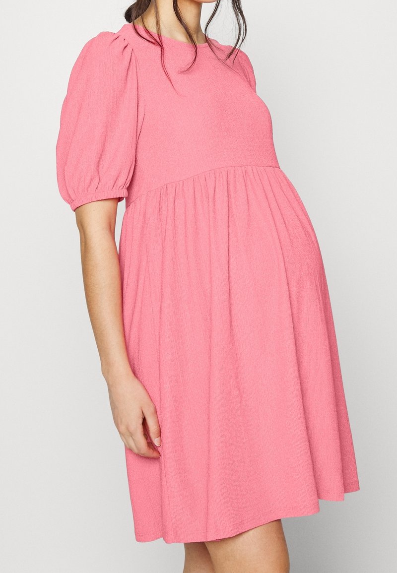 Una donna che indossa un vestito rosa a maniche corte con maniche a sbuffo e vita arricciata, in piedi contro uno sfondo leggero e semplice.