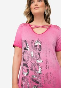 Vaaleanpunainen rento t-paita, jossa on koristeellinen ristikkonokka, lyhyet hihat, paljeteista tehty tekstipainatus sekä pehmeä, hieman teksturoitu kangas.