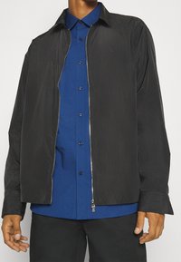 Mand iført en blå skjorte med knapper under en sort jakke med lynlås, lange ærmer og krave, stående mod en ensfarvet baggrund.