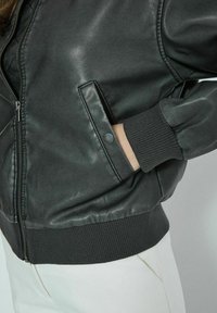 Giubbotto bomber in pelle nera con zip frontale, polsini a coste e tasche laterali con dettagli a pattina. Texture liscia e design aderente.