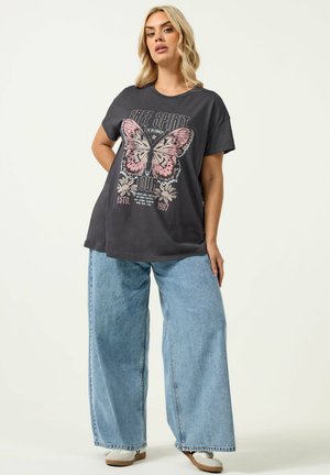 Kvinde med blondt hår iført en stor sort T-shirt med en lyserød sommerfugl og lyseblå jeans med vide ben, stående med den ene hånd på hoften.