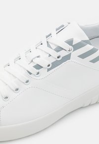 White sneakers with a textured leather upper, featuring gray accents and stripes, flat white laces, and a smooth rubber sole.