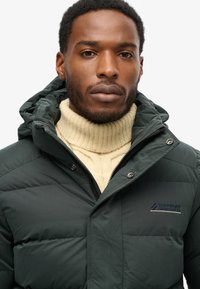 Mørkegrøn pufferjakke med hætte, der har en lynlåslukning og trykknapper. Bæres over en lys cremefarvet strikket turtleneck. Logo på brystet.