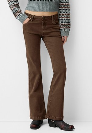 Person iført brune bootcut-bukser, en grå mønstret cropped sweater og sorte støvler med hæl, stående mod en ensfarvet hvid baggrund.