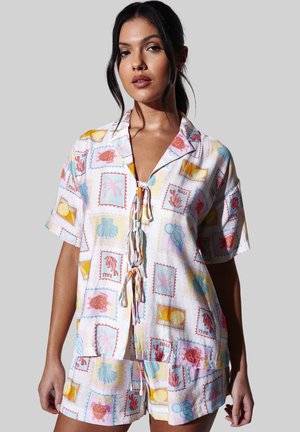 Vrouw draagt een kleurrijk, kort mouwens overhemd met knopen en bijpassende korte broek met tropische stempelpatronen op een lichte achtergrond.