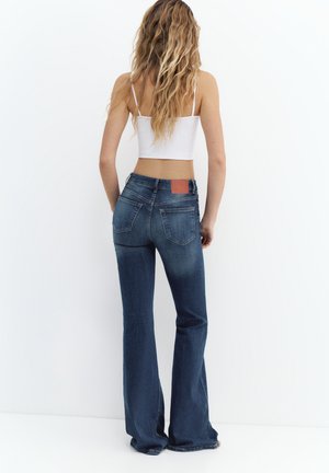 Žena s dugom valovitom kosom, odjevena u bijeli kratki top i tamnoplave traperice visokog struka s proširenim nogavicama, stojeći ispred obične bijele pozadine.