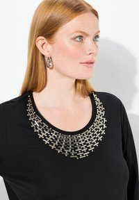 Zwarte top met een bewerkte zilveren kraag, lange mouwen en een gladde textuur. Model draagt hangerogen die de look verfraaien.