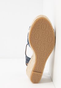 Sandale compensée avec semelle en jute tissée, brides bleu marine et semelle extérieure en caoutchouc à motif, reposant contre un mur blanc en angle.