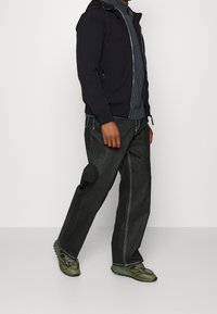 Mørk marineblå lynlåstrøje, grå sweater og mørke denimbukser med hvide syninger. Iført grønne sportssko med strukturerede såler.