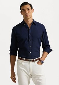 Cămașă button-up de culoare albastru marin, din bumbac, cu două buzunare frontale, mâneci lungi împăturite, asortată cu blugi albi și o curea maro.