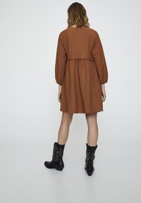 Braunes, strukturiertes Kleid mit langen Ärmeln und gerafftem Taillenzug, kombiniert mit schwarzen Cowboy-Stiefeln mit komplizierter silberner Stickerei.