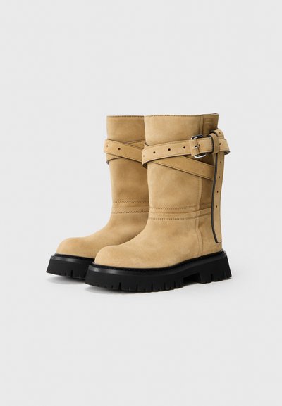 Para beżowych zamszowych botków do połowy łydki z czarnymi, masywnymi podeszwami i paskami z klamrami owiniętymi wokół górnej części.