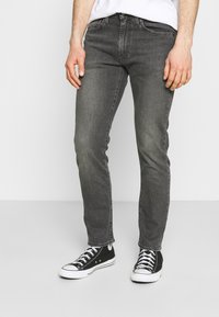Jeans de mezclilla gris de corte ajustado, con cinco bolsillos, una cinturilla clásica y detalles de desgaste sutiles. Combinados con zapatillas de lona negras.