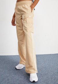 Μπεζ cargo παντελόνια με ελαστική μέση, πλαϊνές τσέπες και τρεις λευκές ρίγες κατά μήκος των εξωτερικών ποδιών; κατασκευασμένα από απαλό, ελαφρύ ύφασμα.