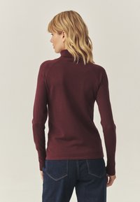 Pulover burgundy cu guler rolă, cu un design ajustat, mâneci lungi și guler cu dungi, asortat cu blugi de denim închis. Textură netedă.