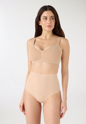 Kobieta o długich, brązowych włosach, ubrana w beżowy, bezszwowy biustonosz i wysokie majtki, patrząca lekko w lewo.