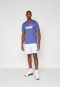 Violeta T-krekls ar lielu baltu "PUMA" logotipu un rozā akcentu, apvienots ar baltām šortiem ar melnu detaļu, valkāts ar baltām sporta apaviem.