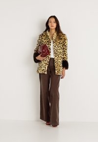 Casaco de pelúcia imitação de leopardo com detalhes pretos, combinado com uma blusa branca e calças largas castanhas. Segurando uma carteira em maroon.