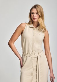 Beige ärmlös klänning med krage, framknappring och knuten midja. Tillverkad av mjuk tyg med en avslappnad passform.