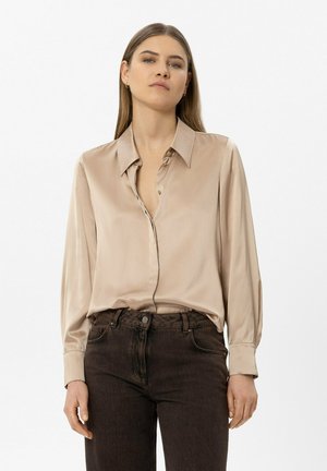 Frau mit glattem Haar, die eine beigen Satinbluse in dunkelbraune hoch taillierte Jeans gesteckt trägt, vor einem einfarbigen Hintergrund.