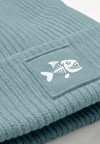 A light blue knit beanie featuring a textured pattern and a rectangular patch with a white fish graphic.