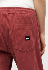 Nærbilde av en person som har på seg røde kordfløyelsbukser med elastisk midjebånd og en baklomme med en svart-hvitt logo-merkelapp.